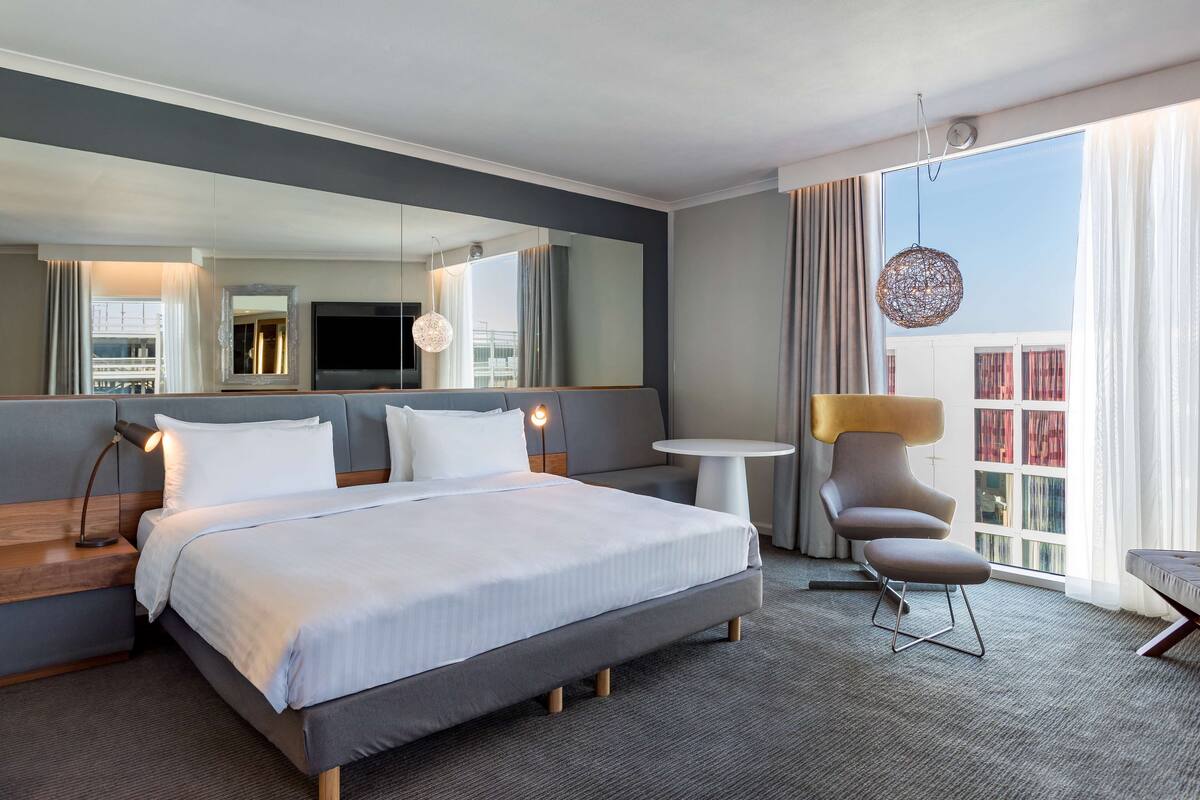 A modern hotel room with a large bed, a small round table, a comfortable armchair, a large mirror, and a view of the city through the window.