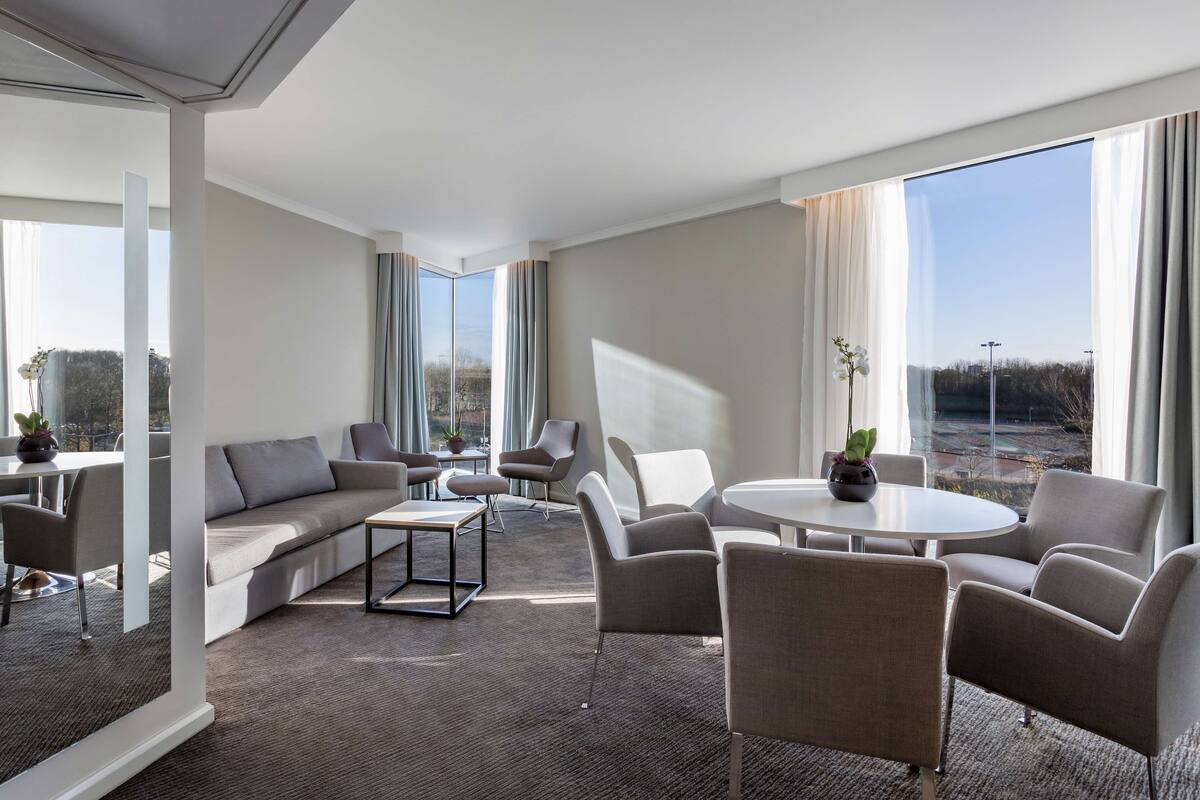 A modern hotel room with a large window, a sofa, a dining table, and several chairs.