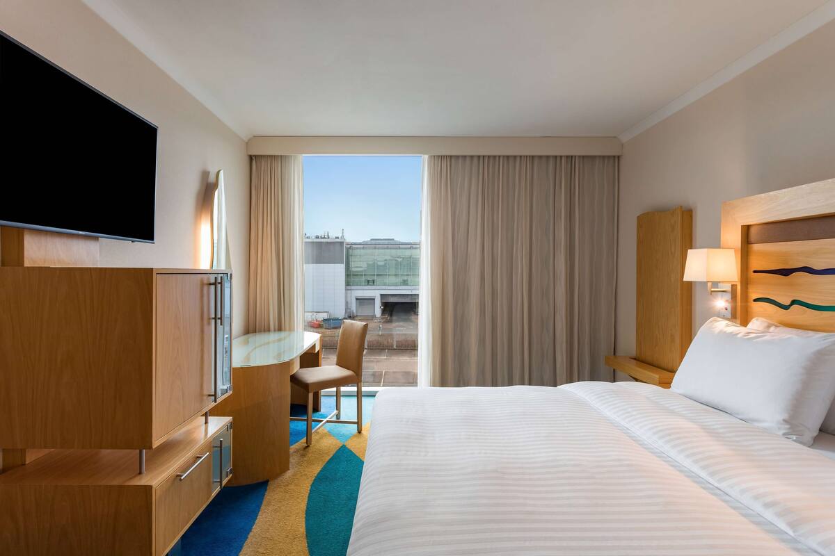 A hotel room with a bed, a desk, a chair, a television, and a view of the outside.
