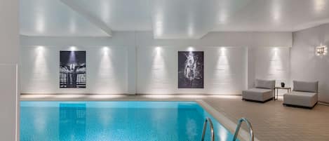 A modern indoor swimming pool with clear water, surrounded by white walls and minimalist furniture.