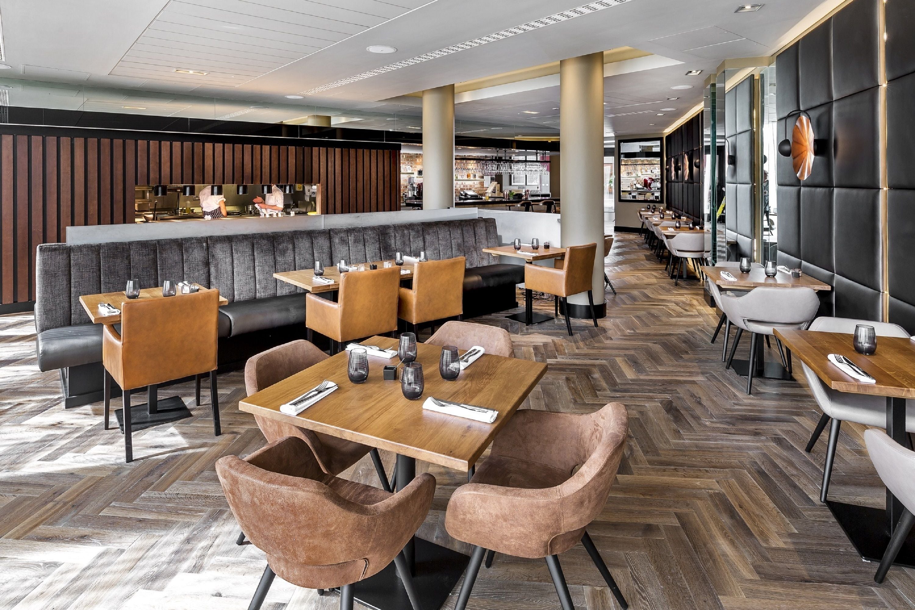 En modern restaurang med ett parkettgolv i fiskbensmönster, läderbeklädda sittplatser och träbord med vadderade stolar.