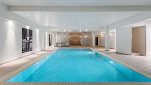 Una piscina interior moderna con superficie de agua azul cristalina, rodeada de paredes blancas y suelo de baldosas.