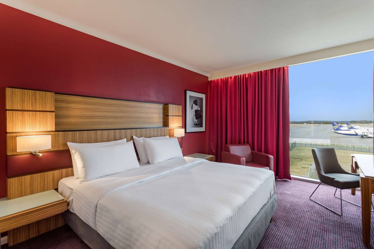 A hotel room with a large bed, a desk, a chair, a red curtain, and a view of an airport.