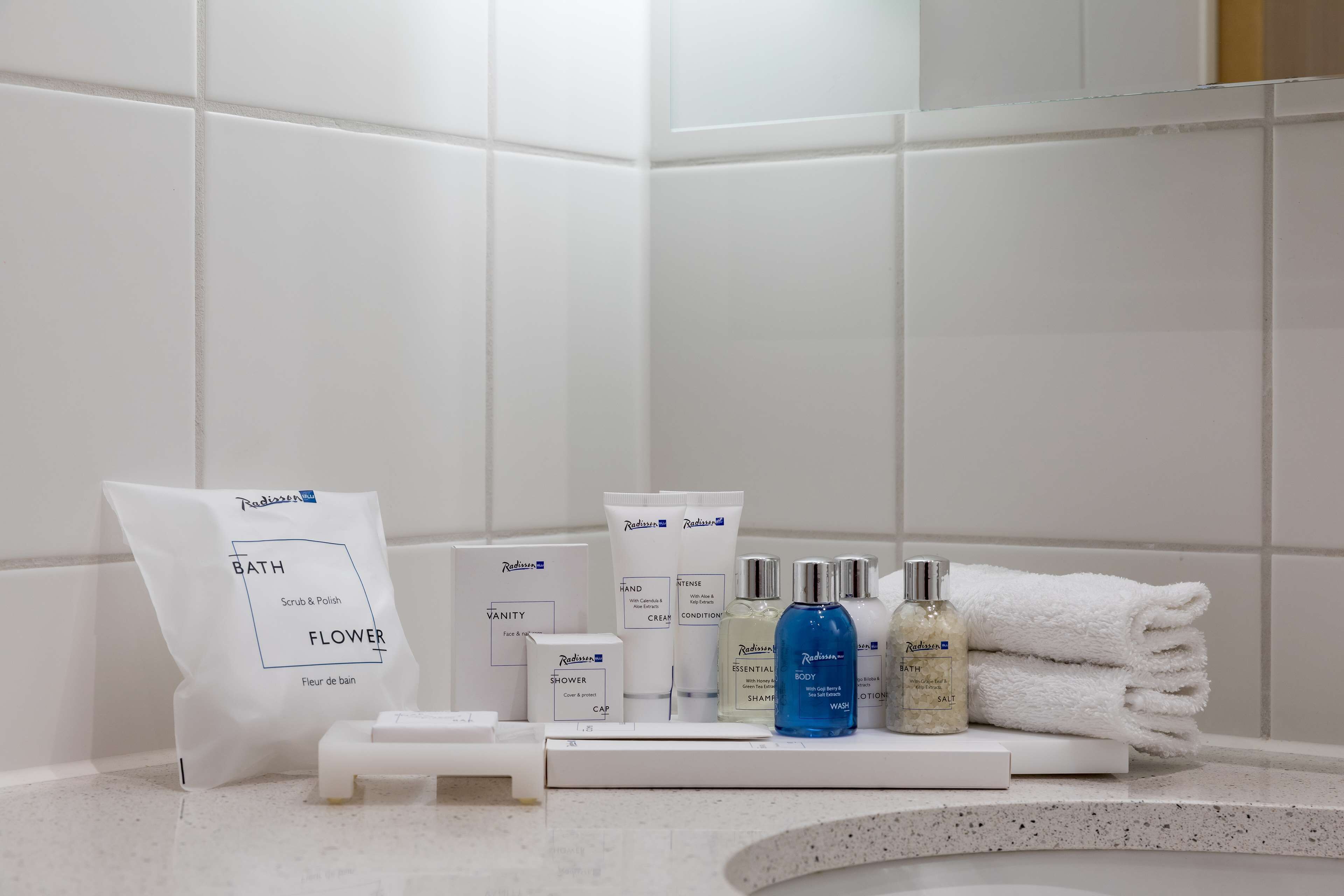 A bathroom with white tiles, a white towel stack, and various toiletries including a bottle of blue liquid, a bottle of clear liquid, and a white bag labeled "Bath Scrub & Polish."