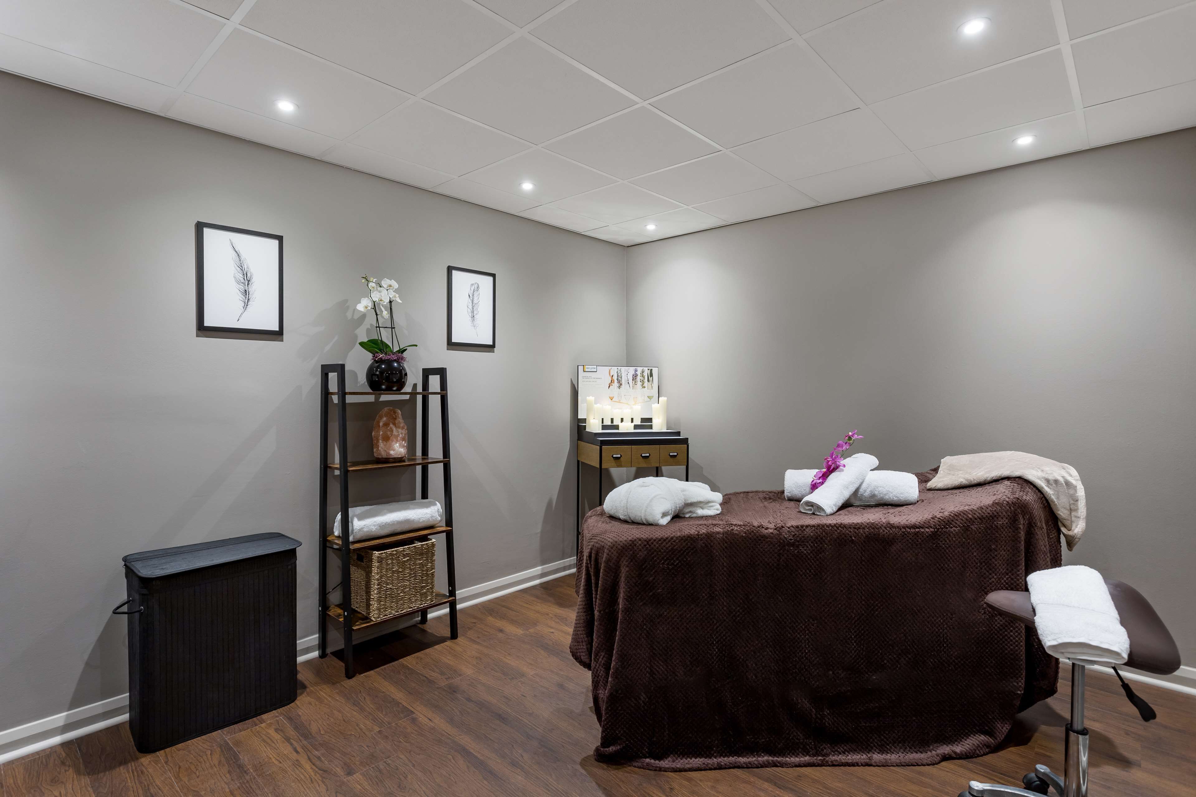 Ein Spa-Raum mit Massageliege, einem Regal mit Handtüchern, einem kleinen Tisch mit Lampe und gerahmten Kunstwerken an der Wand.