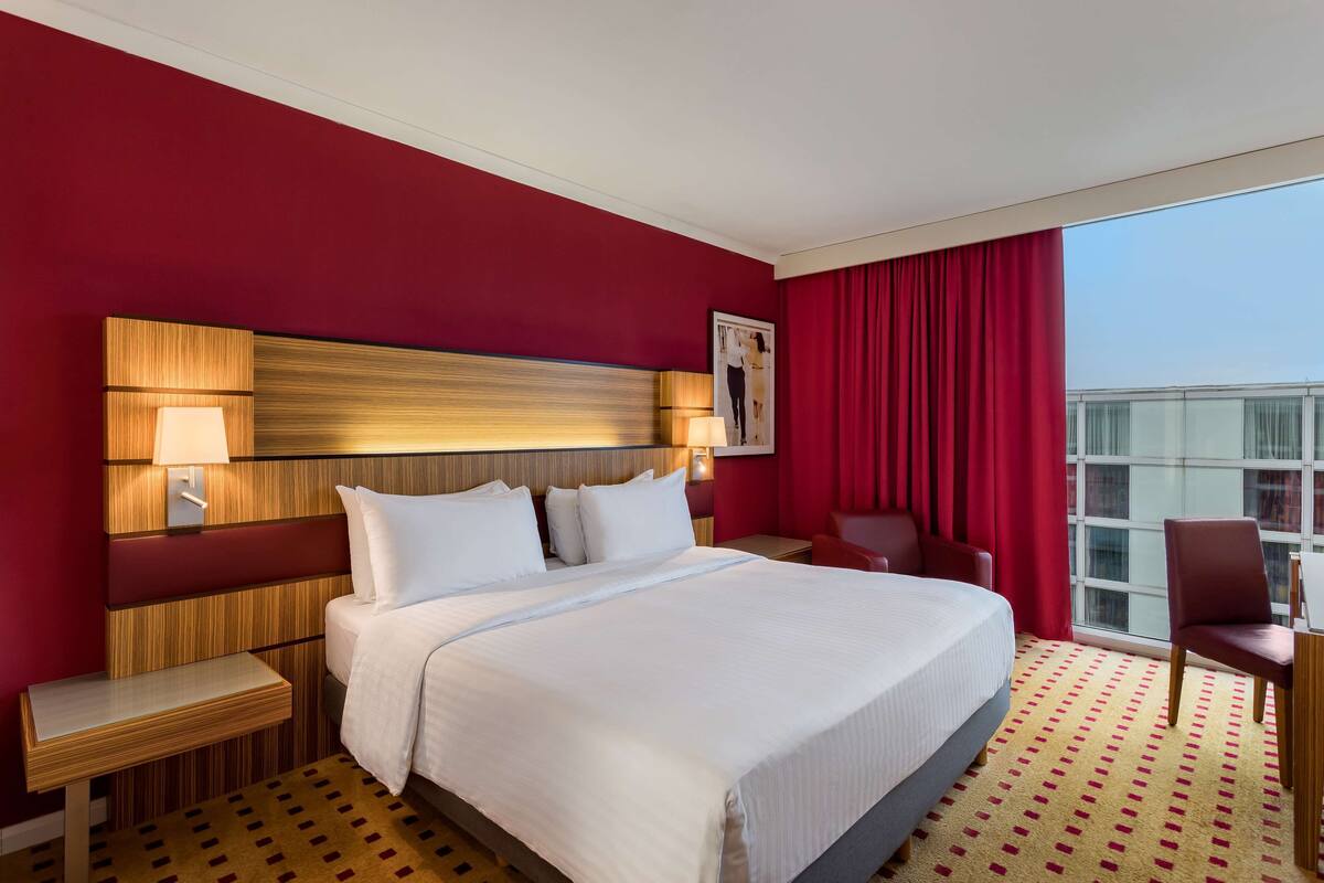 A hotel room with a large bed, a wooden headboard, a nightstand, a desk, a chair, and a large window with red curtains.