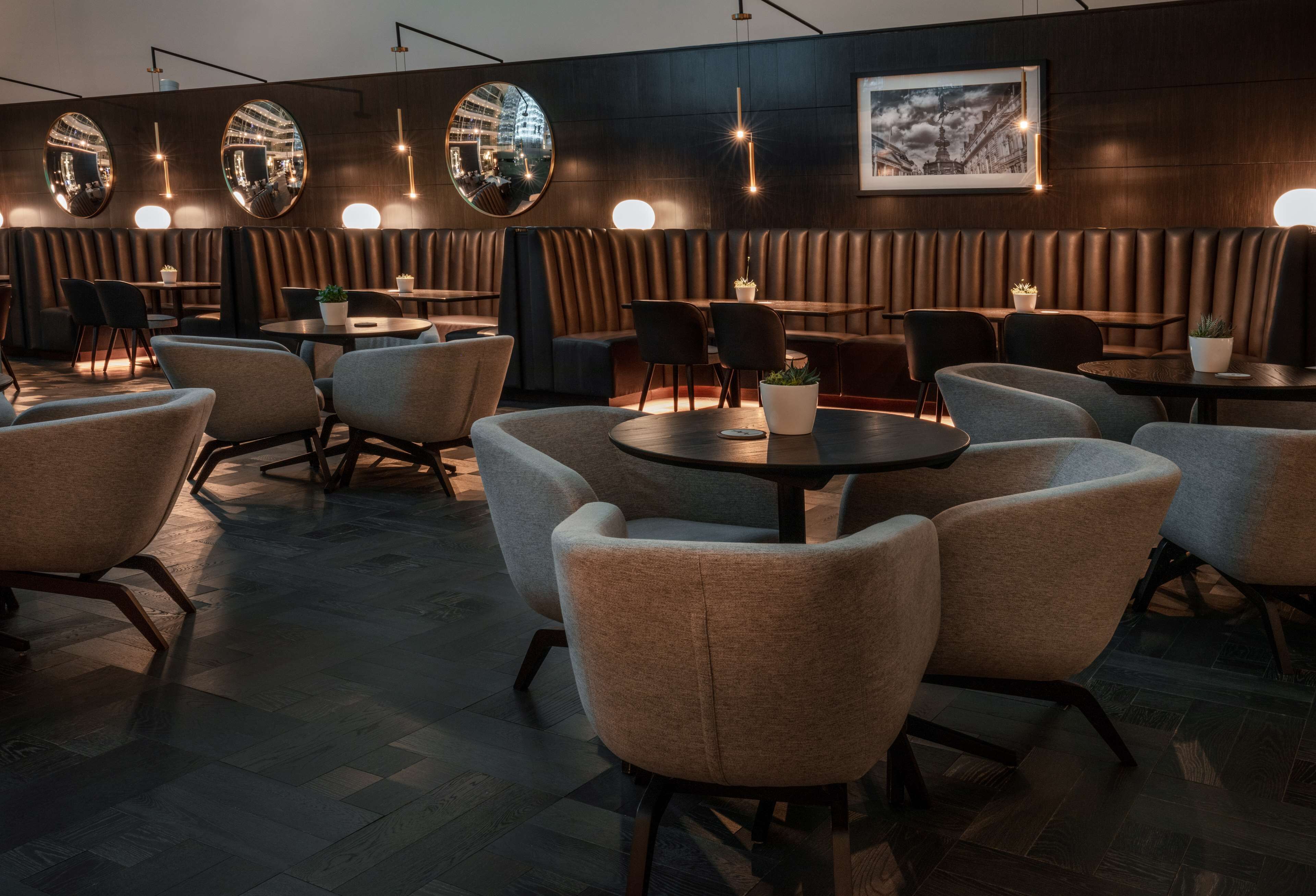A modern restaurant with round tables, cushioned chairs, and a dark wood-paneled wall with mirrors and a framed picture.