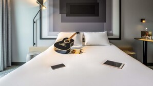 A hotel room with a bed, a guitar, a lamp, and a framed artwork on the wall.
