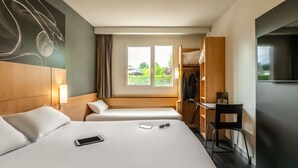 A hotel room with a large bed, a desk, a chair, a TV, and a window with a view of greenery.