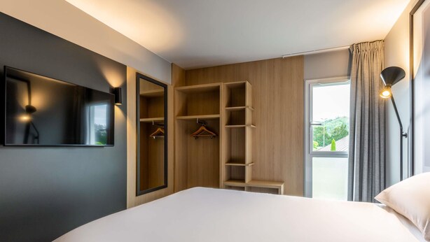 A modern hotel room with a large bed, built-in wardrobe, and a view of greenery outside.