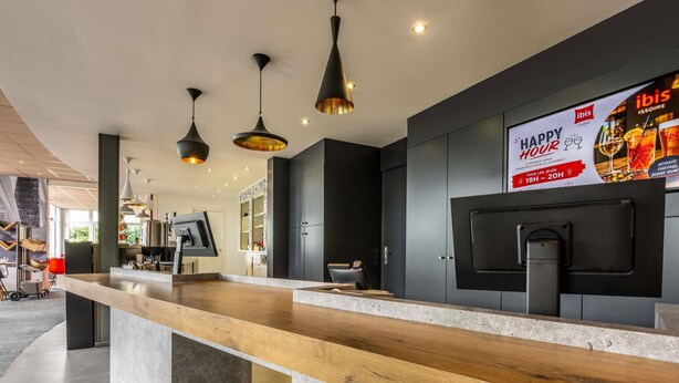 A modern hotel lobby with a wooden reception desk, black pendant lighting, and a large TV screen displaying a 'Happy Hour' advertisement.