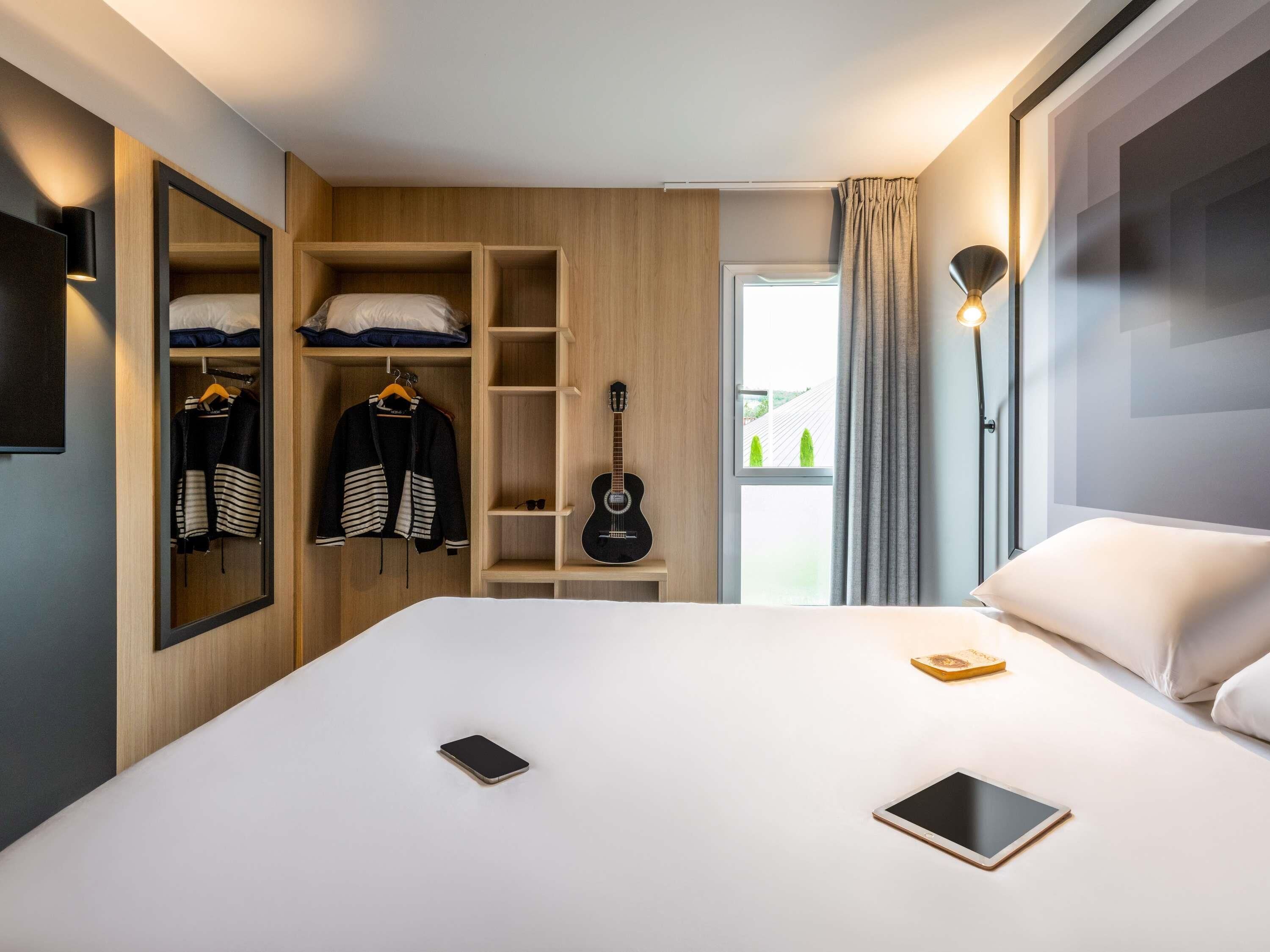 A hotel room with a bed, a wardrobe, a guitar, a bedside lamp, and a window with curtains.
