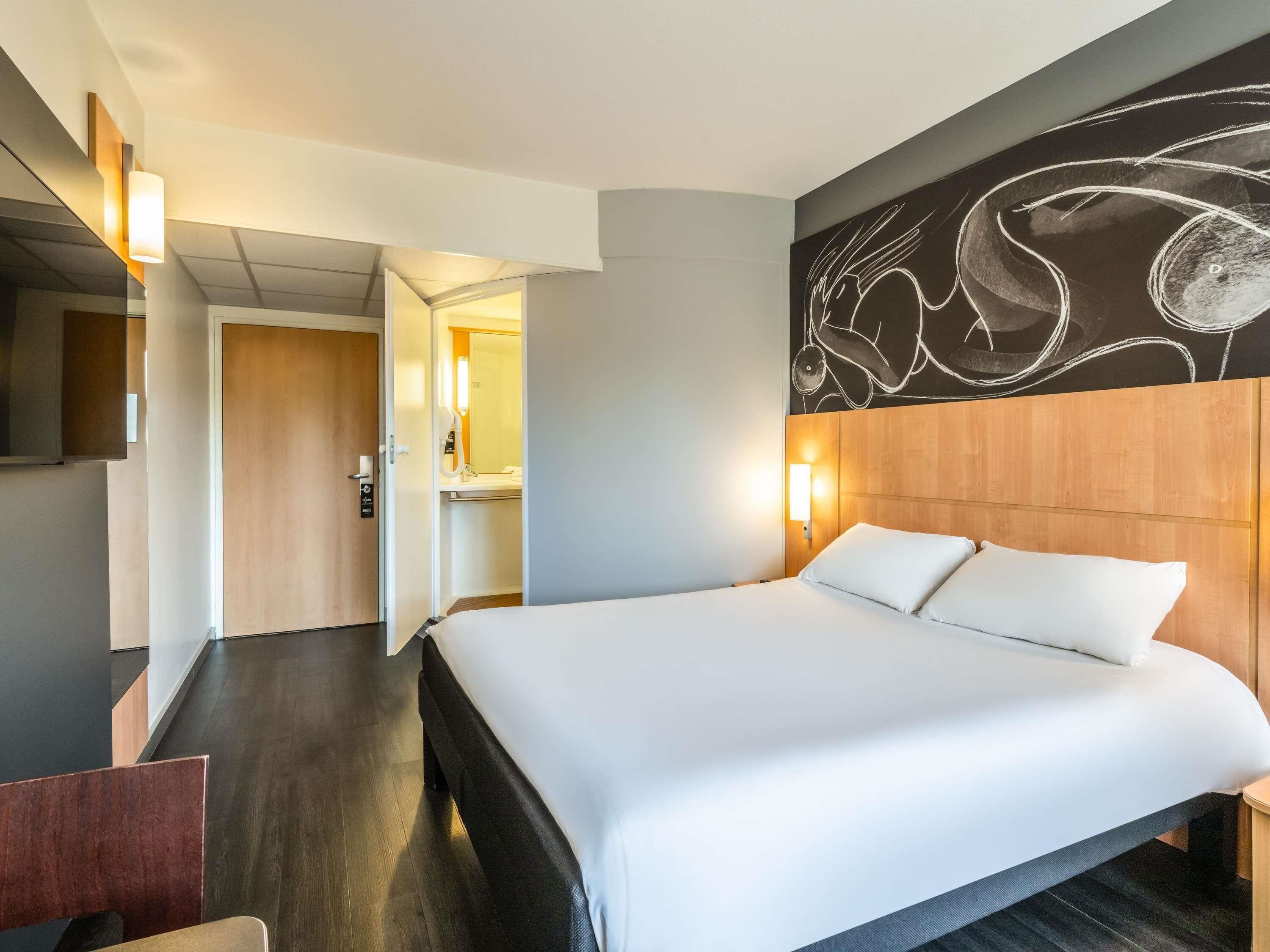 A hotel room with a large bed, a wooden headboard, a wall-mounted artwork, and a door leading to a bathroom.