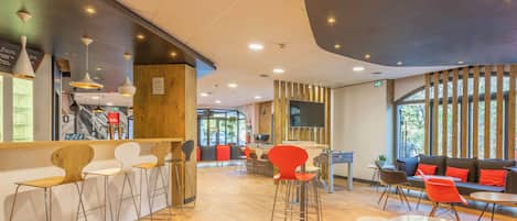 A modern cafe with wooden flooring, bar stools, red chairs, and a seating area with sofas and tables.