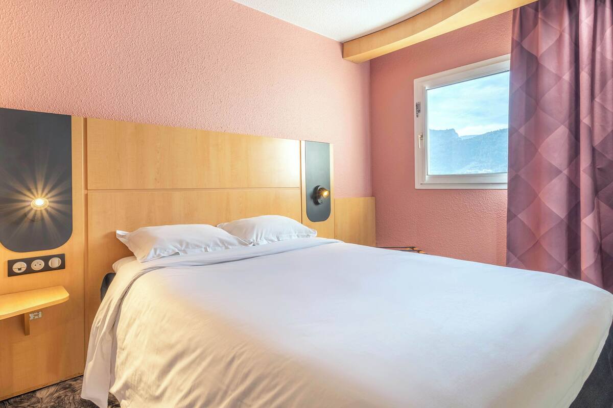 A hotel room with a large bed, a wooden headboard, a nightstand with a lamp, a wall-mounted control panel, and a window with a view of a mountain.