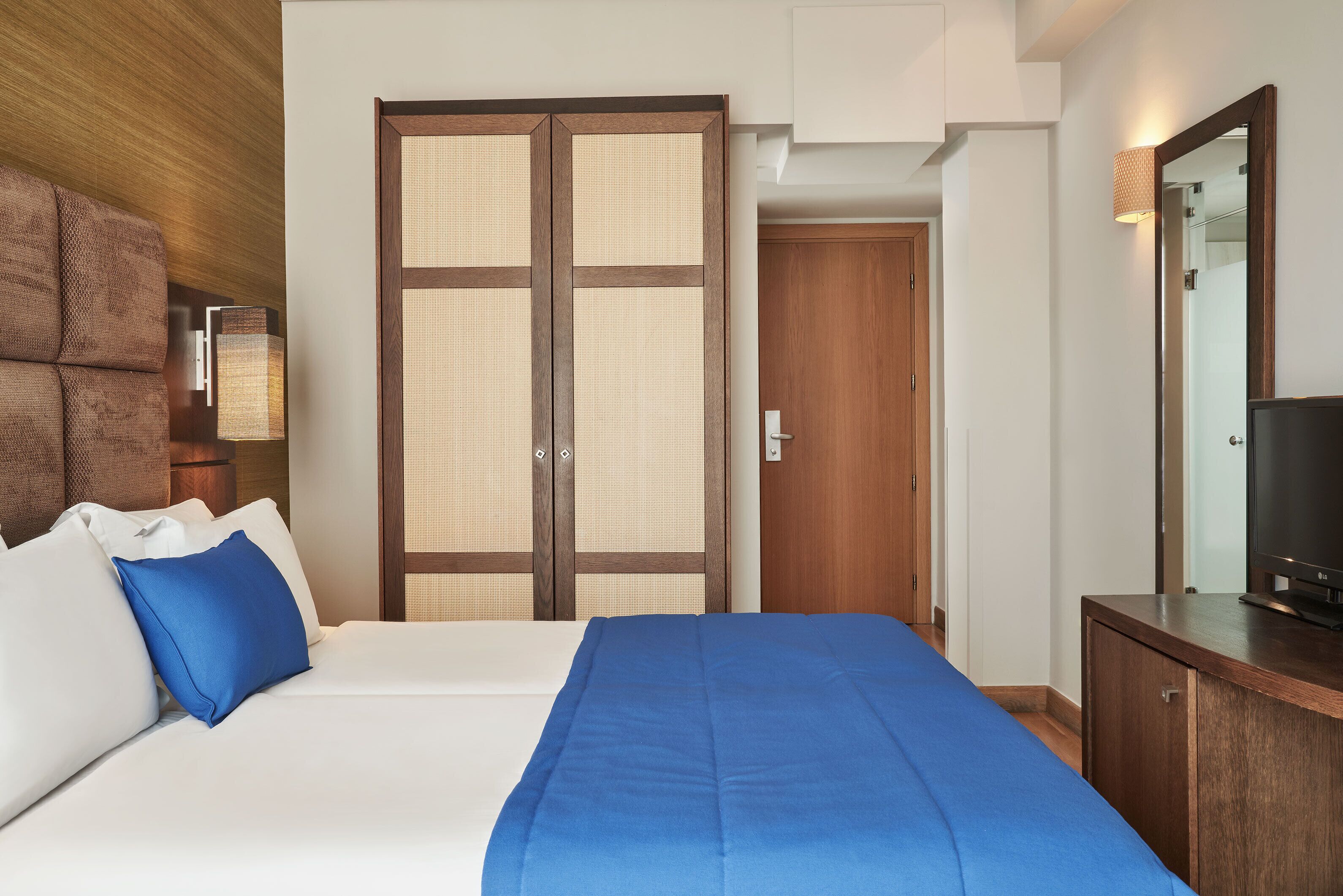 Una habitación de hotel con una cama, un armario de madera, un televisor y un ventanal grande.
