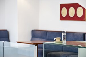 A modern room with a glass table, a white cup, and a glass of water. There is a blue sofa and a wooden table in the background.