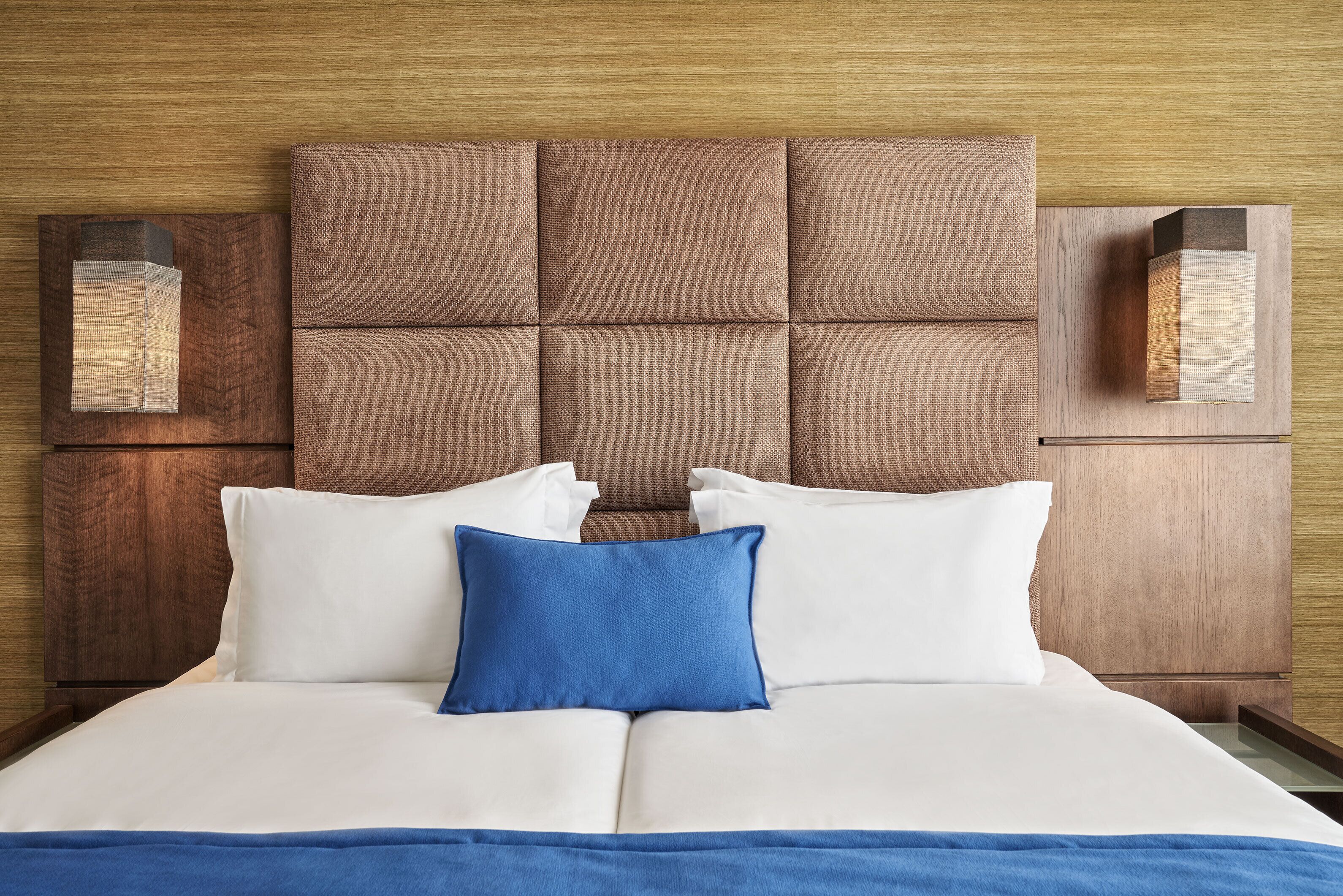 A neatly made bed with white and blue bedding, a tufted headboard, and wooden nightstands with wall sconces.