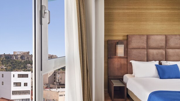 Habitació d'hotel amb un gran finestral, un llit amb roba de llit blanca i blava, un capçal de llit de fusta i vista d'un edifici històric.