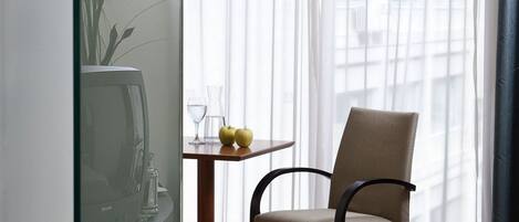 Una habitación de hotel con una cama, una silla, una mesita con un jarrón y dos vasos, y una ventana con cortinas traslúcidas.