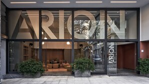 A modern hotel entrance with large glass doors, a seating area inside, and potted plants outside. - Arion Athens Hotel (Athens)