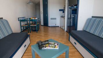 Un espacio habitable compacto con sofá, una pequeña mesa con cámara y revista, zona de comedor con sillas y kitchenette.
