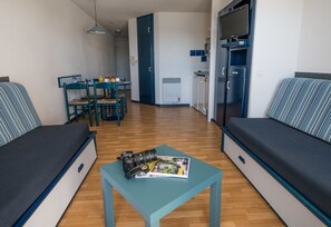 Un espace de vie compact comprenant un canapé, une petite table avec un appareil photo et un magazine, un coin repas avec des chaises et une kitchenette.