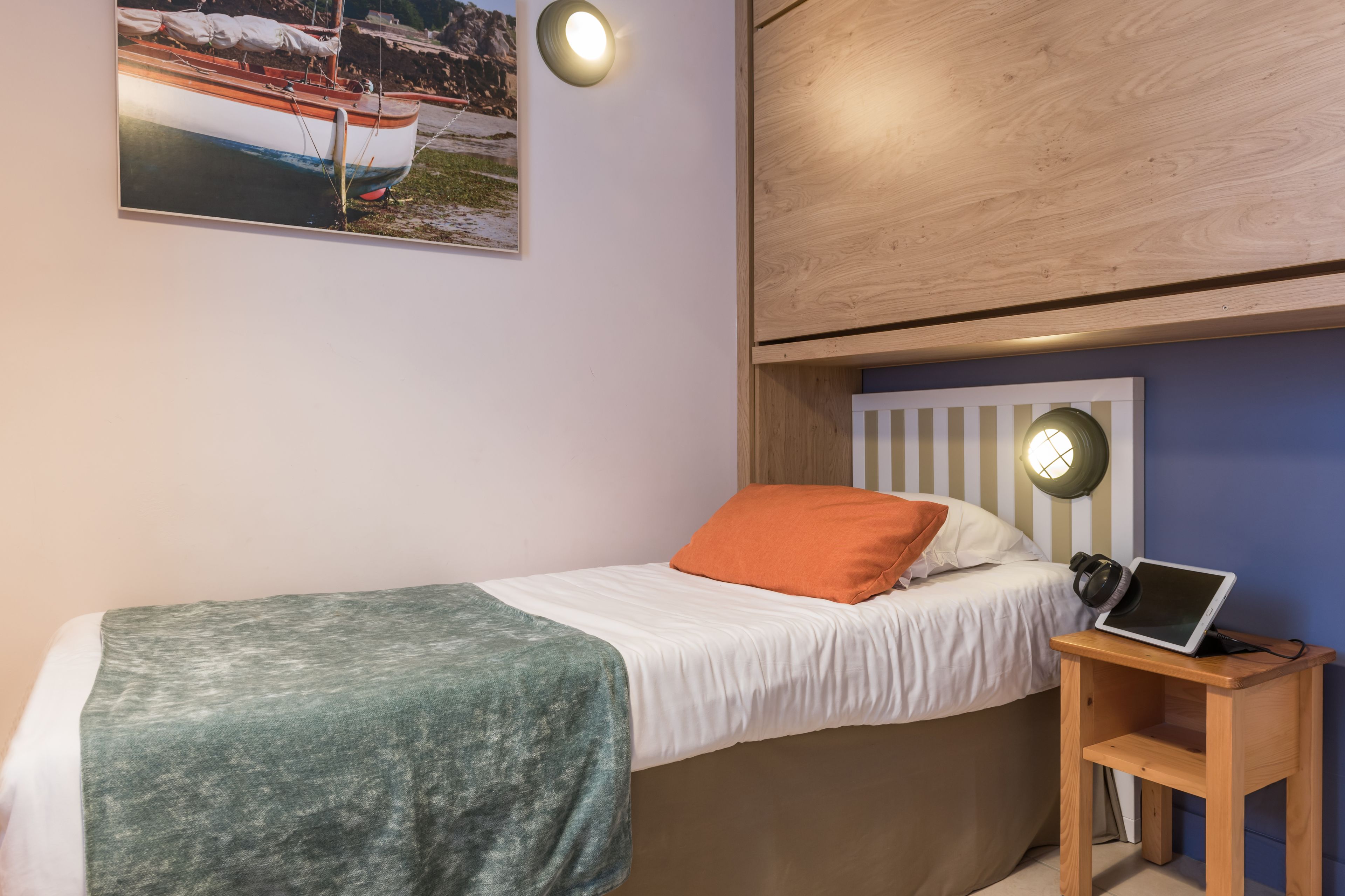 Ein Einzelbett mit einem weiß-orangenen Kopfkissen, einer grünen Decke und einem Laptop auf einem Nachttisch aus Holz.