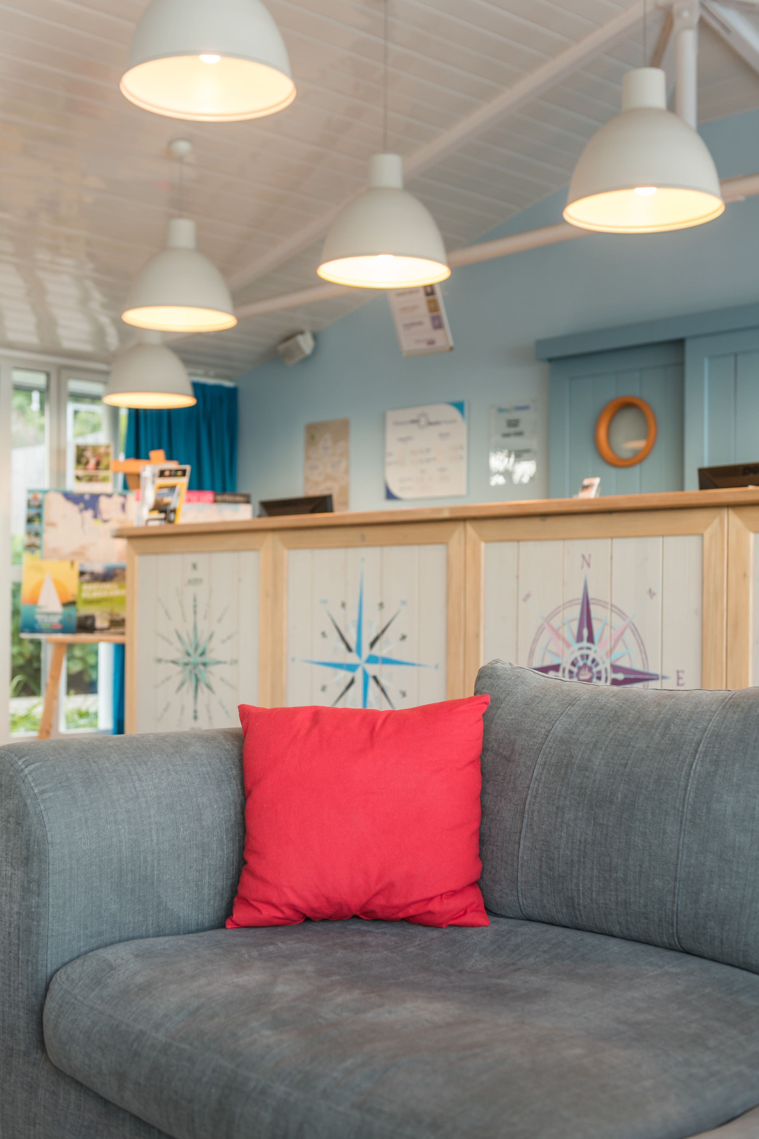 Un coin salon accueillant avec un canapé gris et un coussin rouge, des suspensions blanches au plafond et un comptoir en bois orné d’une fresque représentant une boussole au fond.
