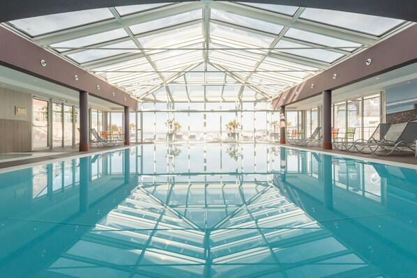 Uma piscina coberta grande com teto de vidro, rodeada por áreas de estar e vista para o exterior.