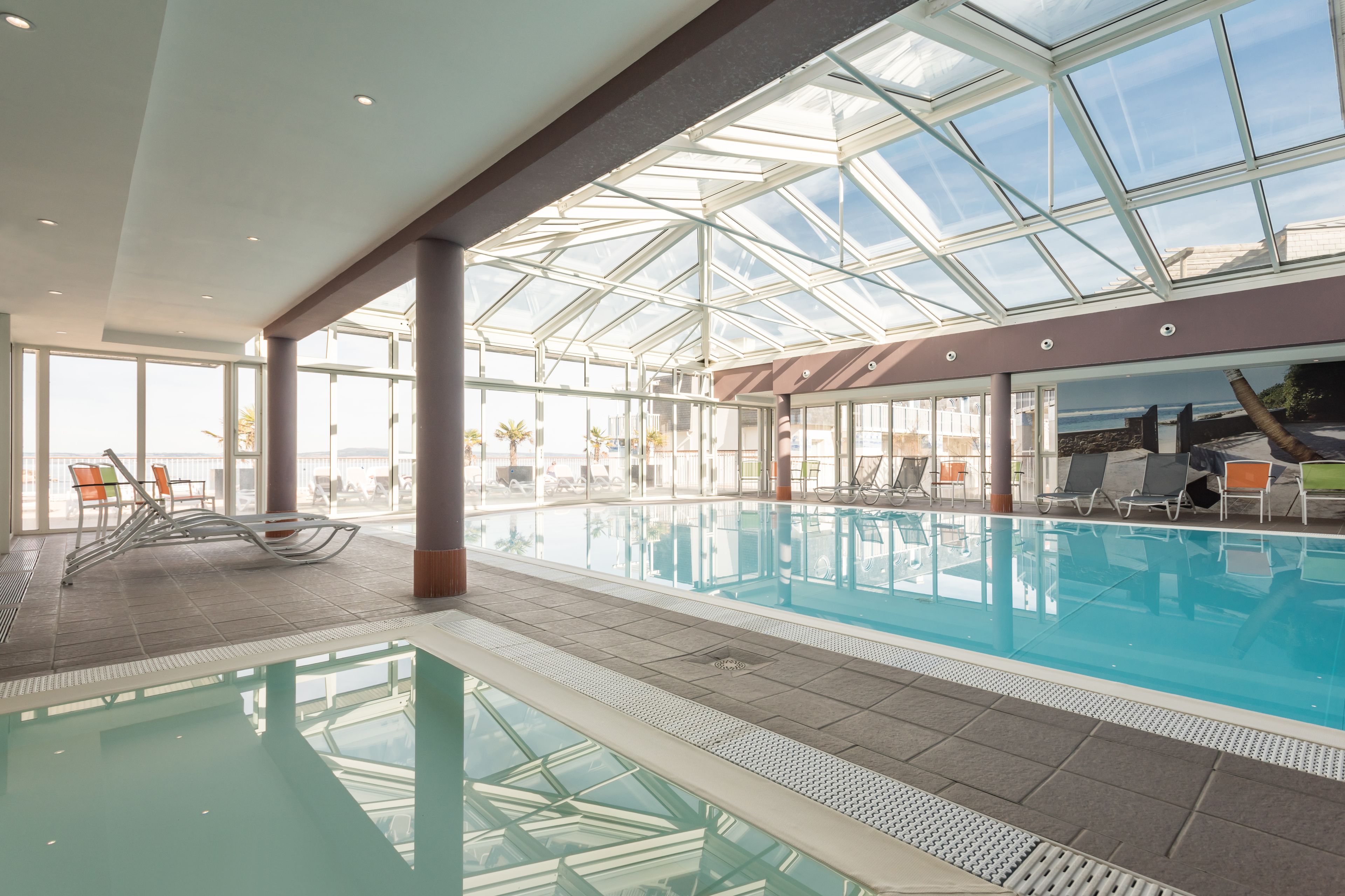 An indoor swimming pool with clear glass ceilings, lounge chairs, and a view of the sea.