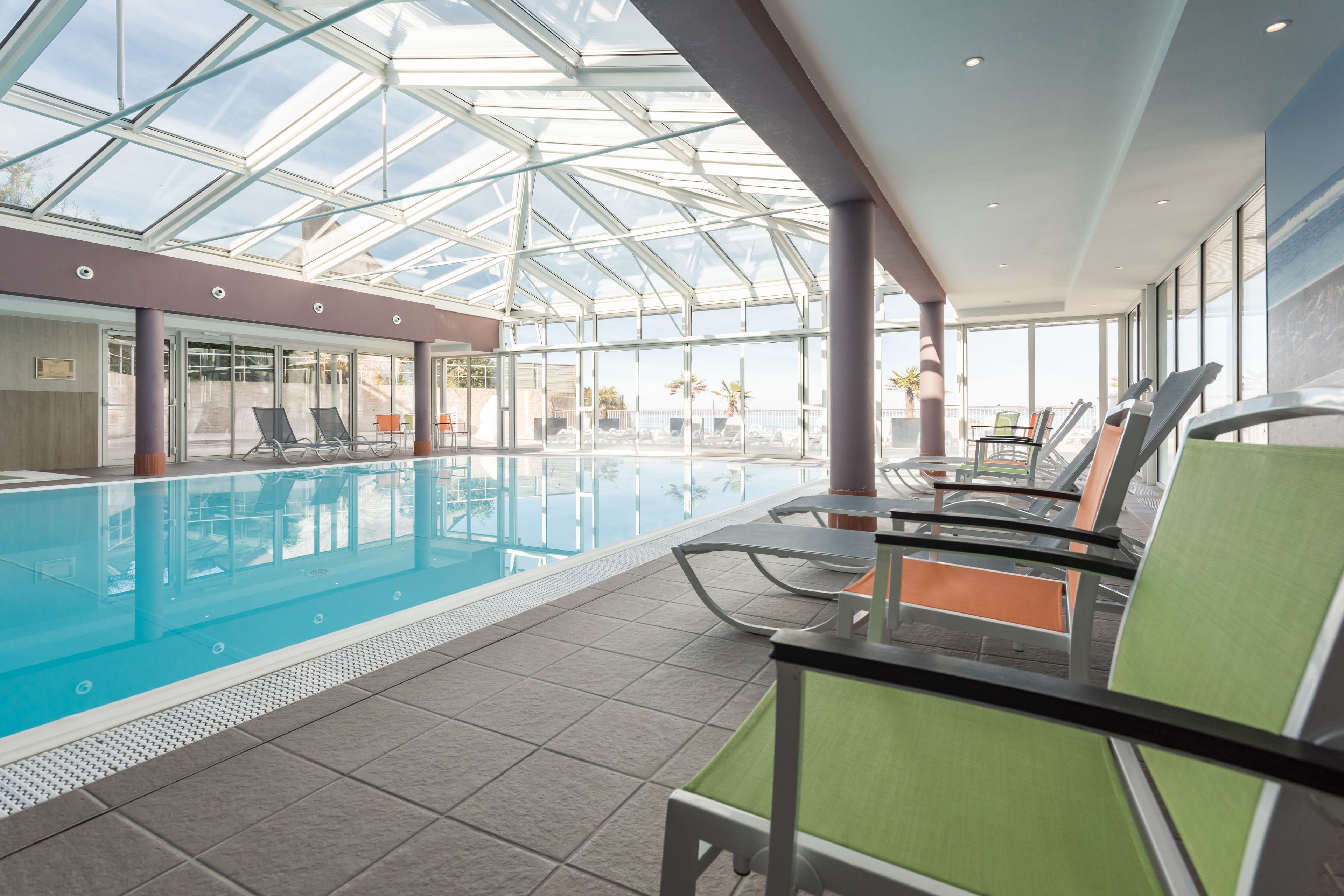 Una piscina interior con techos de vidrio transparente, sillones de descanso y vistas a palmeras.