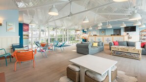 Een ruime binnenruimte met een hoge plafond, verschillende zitmogelijkheden zoals een bank en stoelen, een eettafel en een receptiebalie.