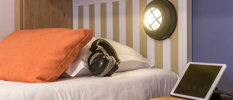 A neatly made bed with white bedding and an orange pillow, a pair of headphones on the bed, and a tablet on a wooden bedside table.