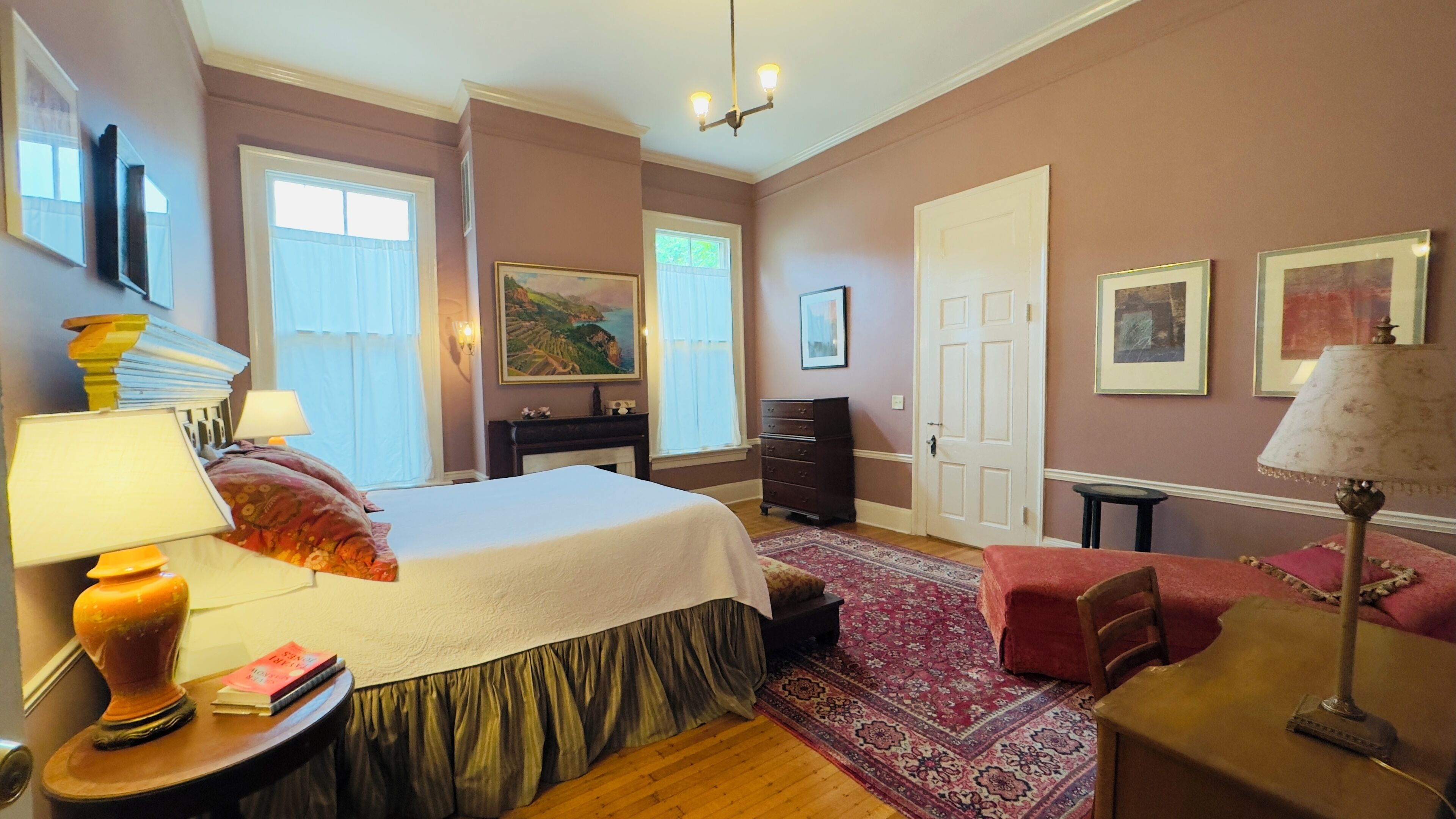 Une chambre à coucher comprenant un lit, des tables de chevet, une commode, une chaise et un bureau. Il y a deux fenêtres avec des rideaux, une porte et un lustre.