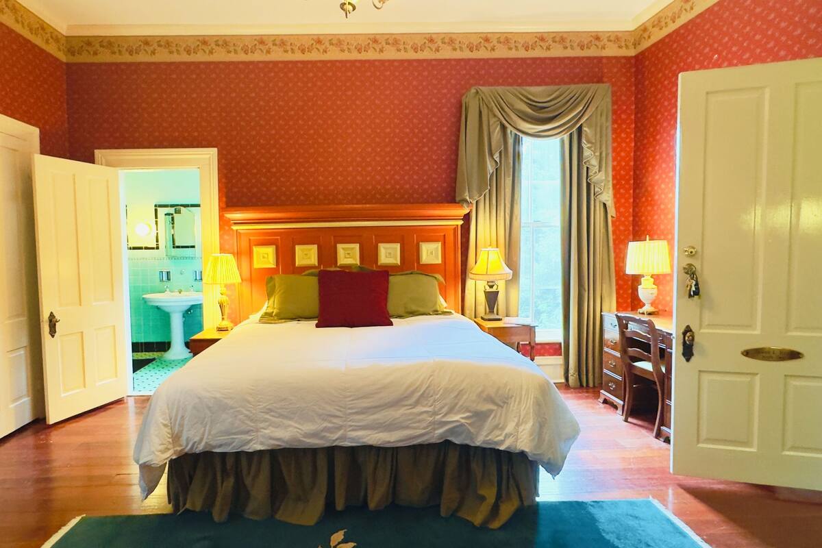 Une chambre d’hôtel avec un grand lit, deux tables de chevet avec des lampes, une tête de lit en bois, une salle de bain visible à travers une porte ouverte et une grande fenêtre avec des rideaux.