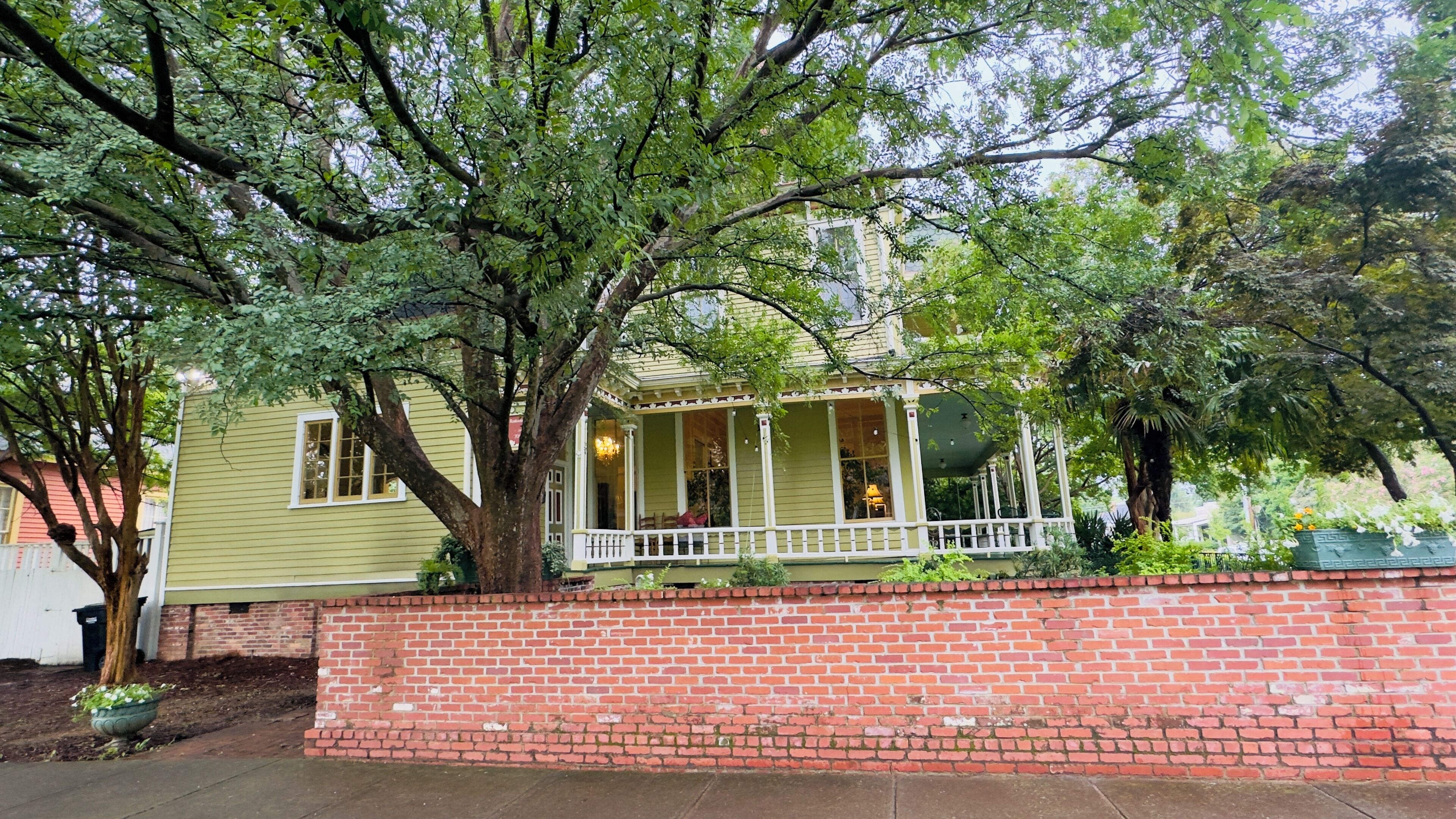 A two-story house with a porch, brick wall, and large tree.