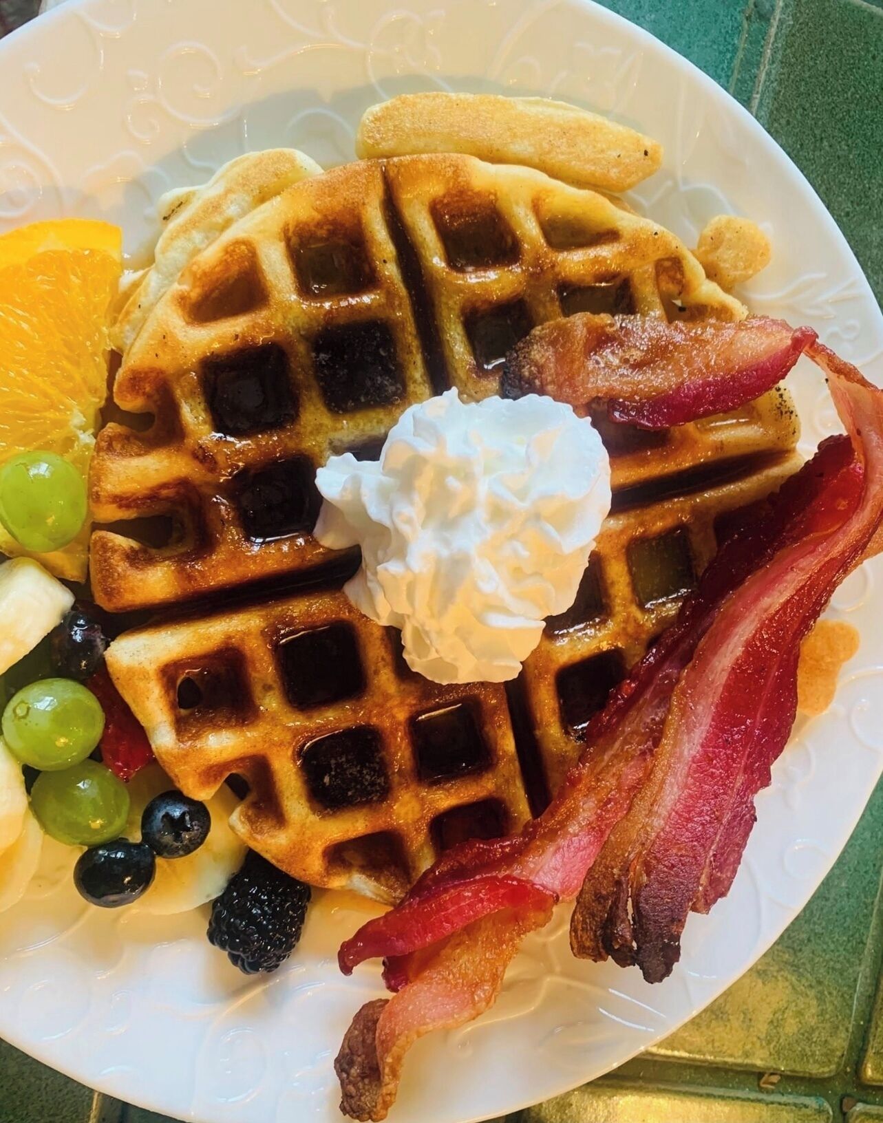 A plate with waffles, bacon, whipped cream, and fruit.