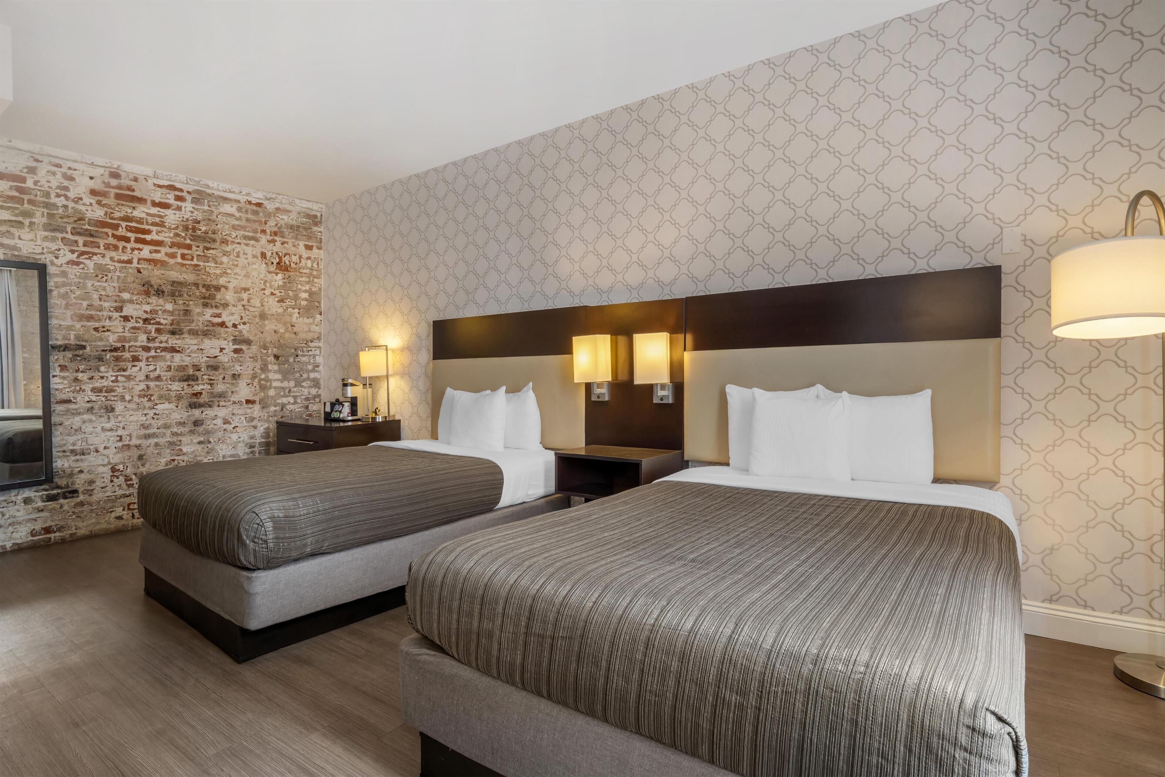 A hotel room with two beds, a brick accent wall, patterned wallpaper, and modern lighting fixtures.