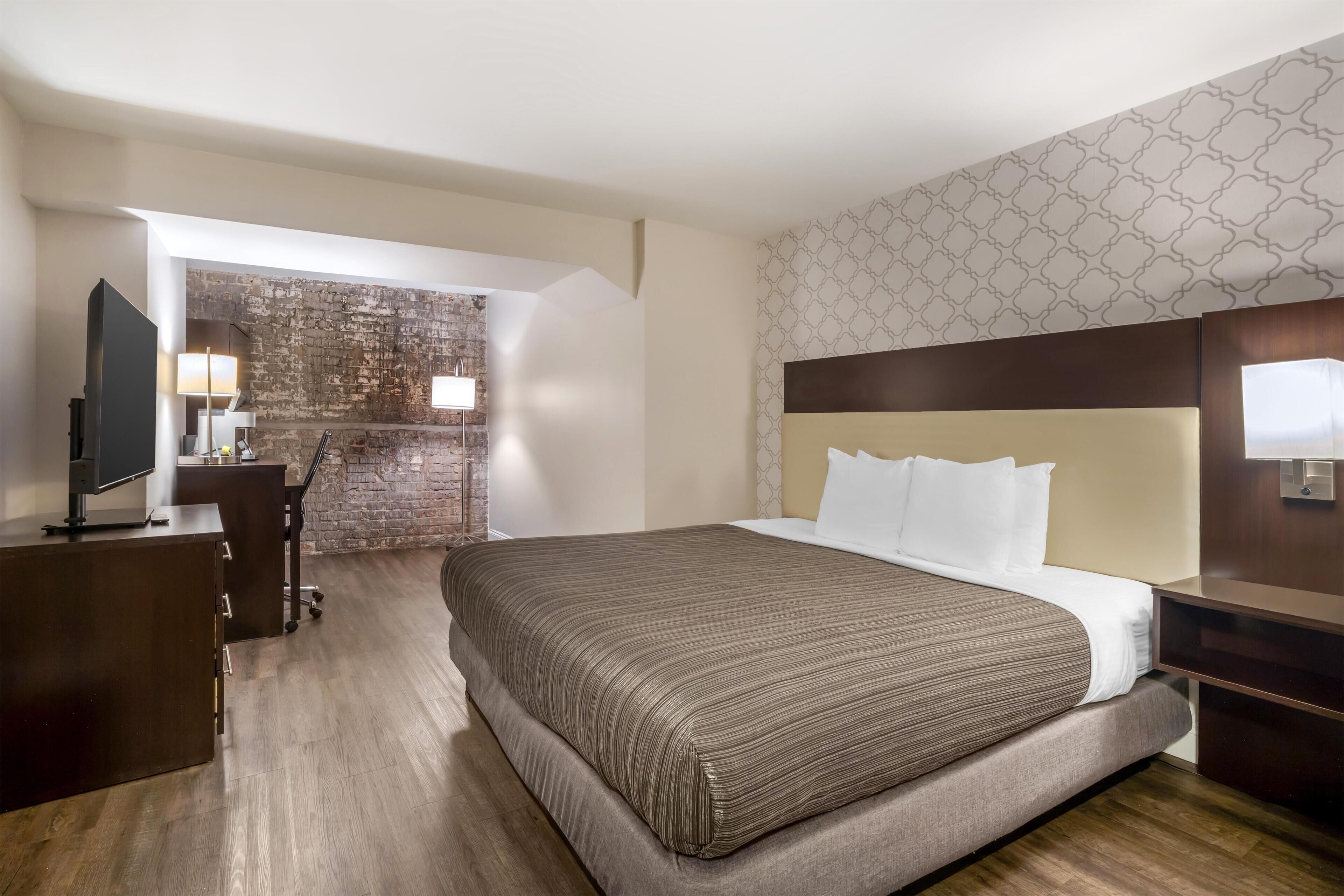 A hotel room with a bed, a desk, a television, and a brick wall.