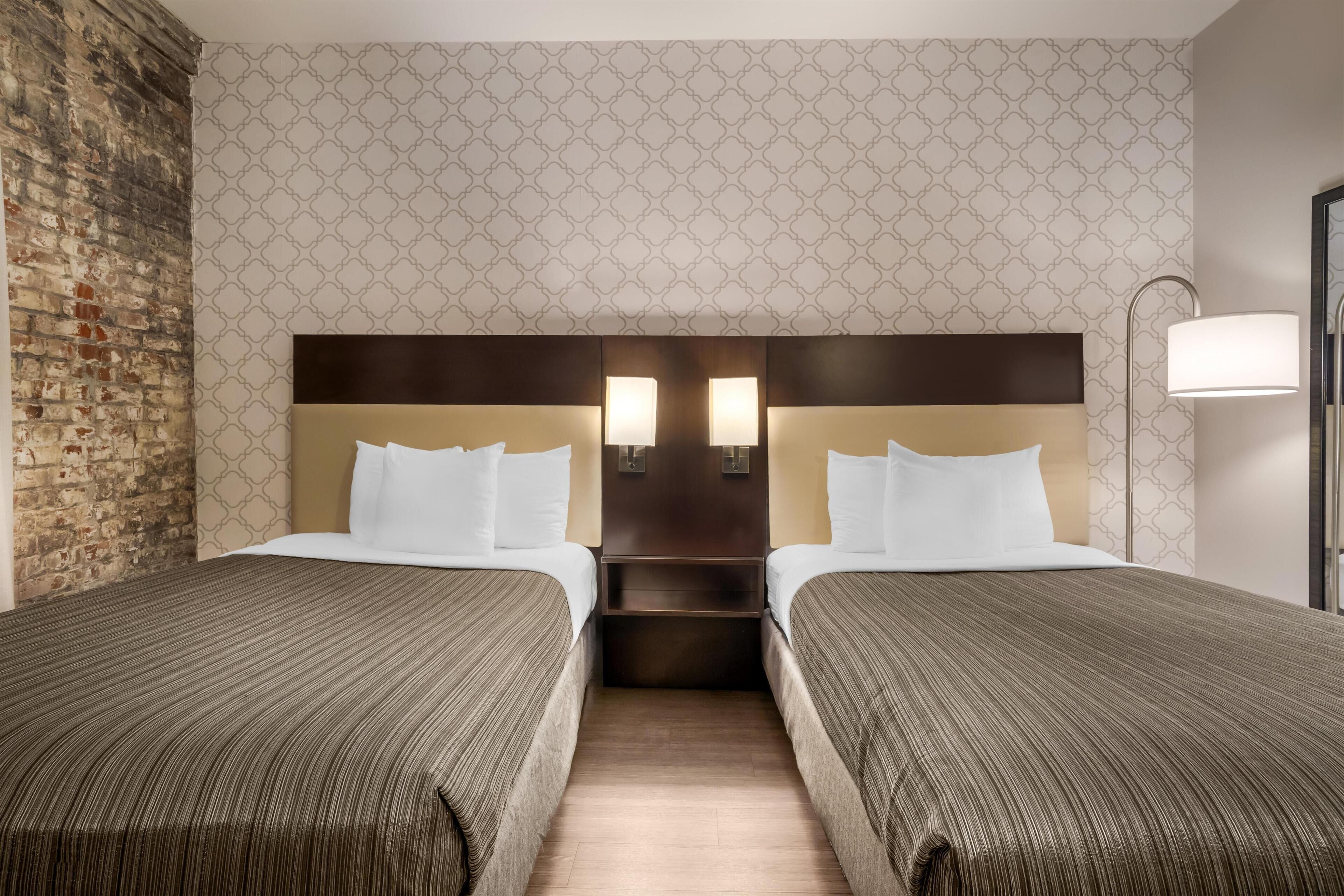 A hotel room with two beds, a brick wall, patterned wallpaper, and a nightstand with lamps.