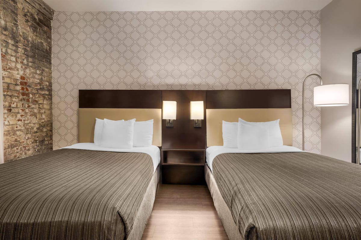 Hotellihuone, jossa on kaksi sänkyä, tiiliseinä, kuvioitu tapetti ja yöpöytä, jossa on lamput.