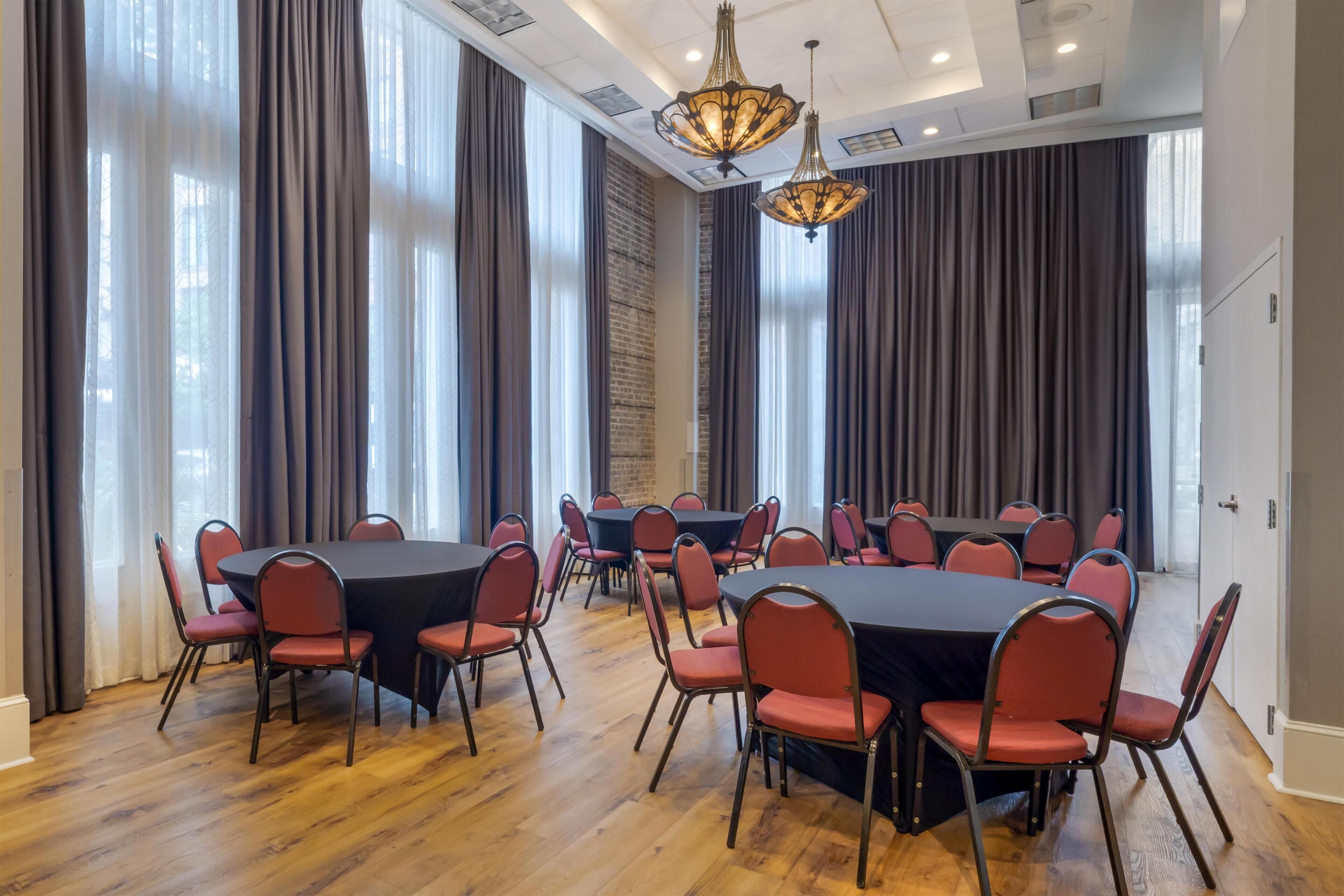 A conference room with multiple tables and chairs, large windows with curtains, and a wooden floor.