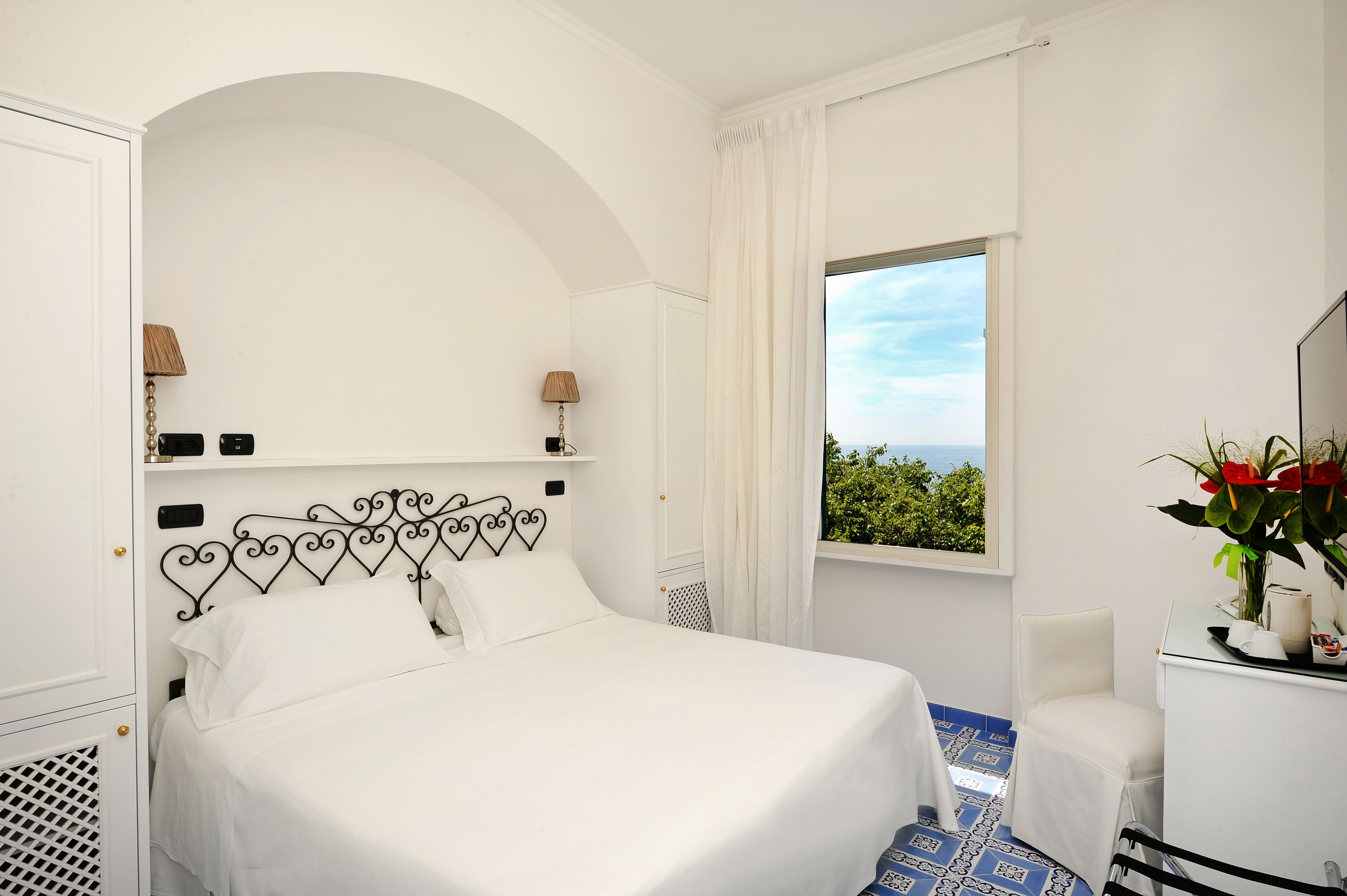 A hotel room with a bed, a desk with a chair, a window with a view, and a decorative plant.