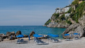A beach with blue umbrellas and lounge chairs, rocky coastline, and buildings on a hillside. - Hotel Aurora (Amalfi)