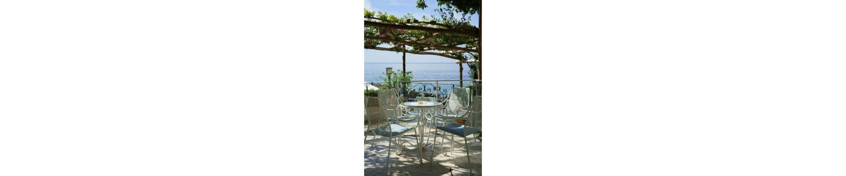 An outdoor seating area with metal chairs and a table, under a pergola with climbing plants, overlooking a body of water.