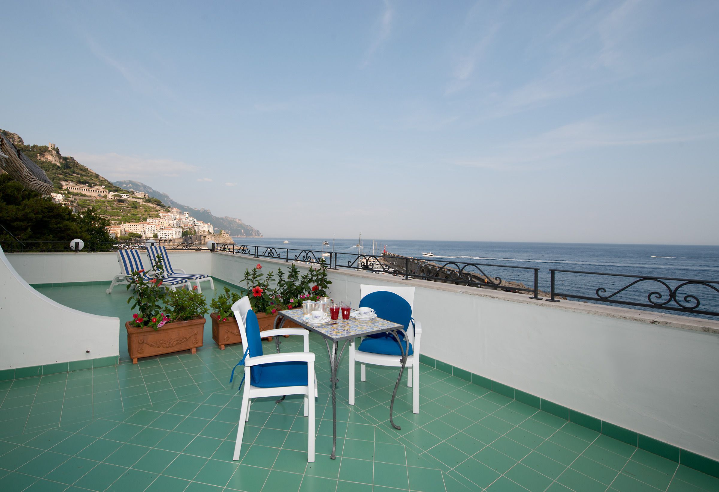 Um terraço com mesa e cadeiras, plantas em vasos e vista para o mar e edifícios.