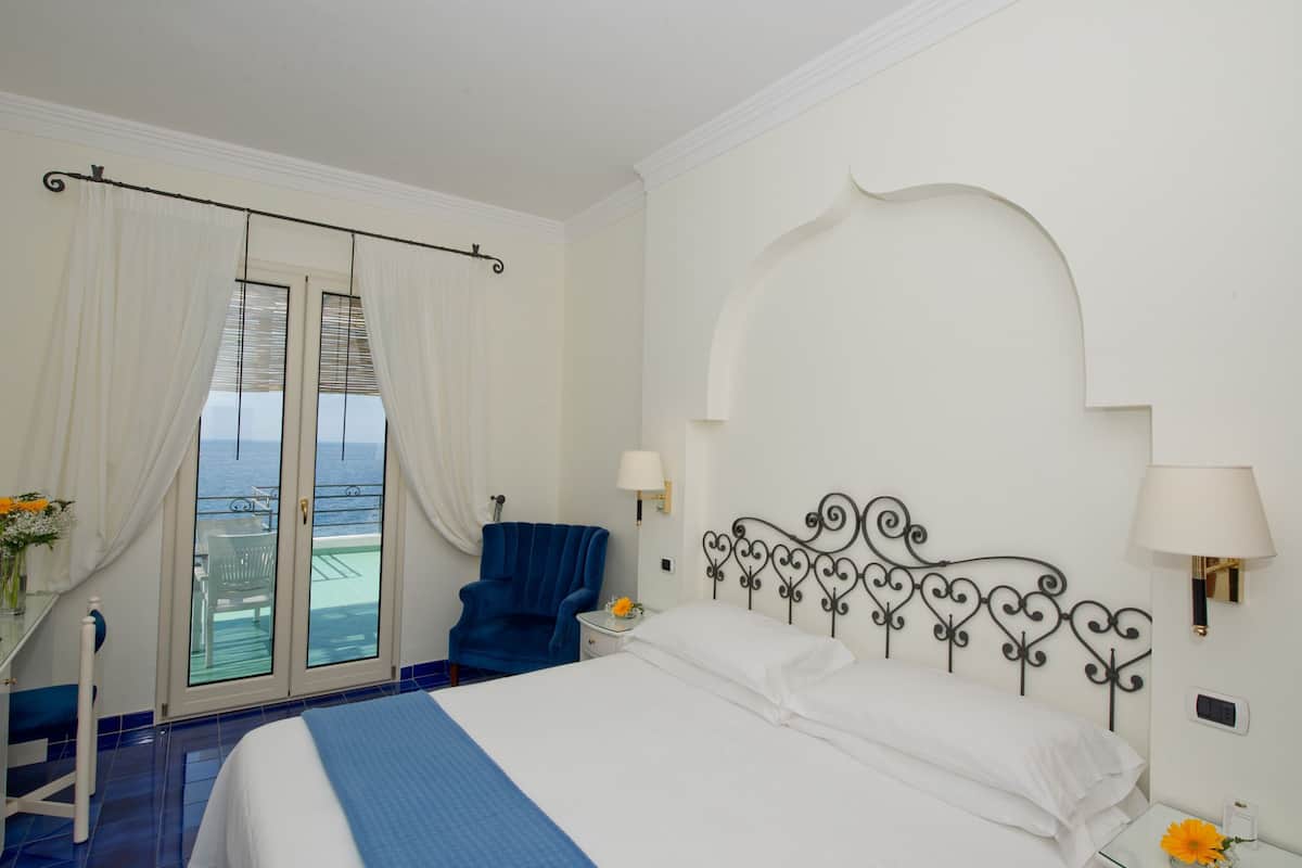 A hotel room with a bed, a chair, a table with a vase, and a view of the sea through a glass door.