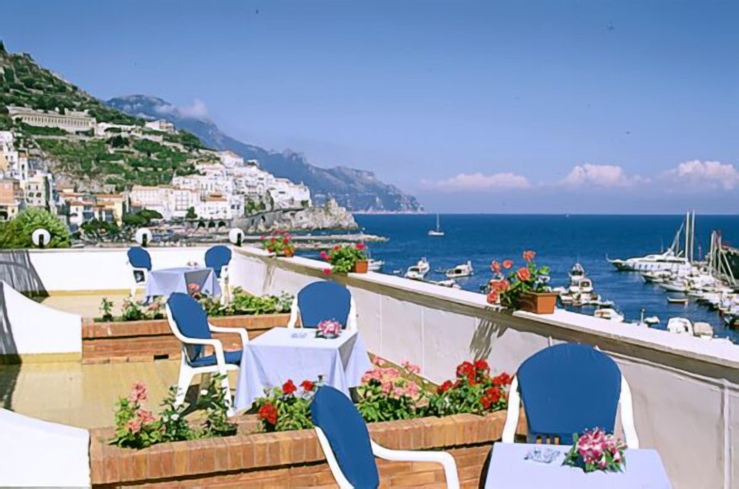 A terrace with tables and chairs overlooking a marina and coastal town.