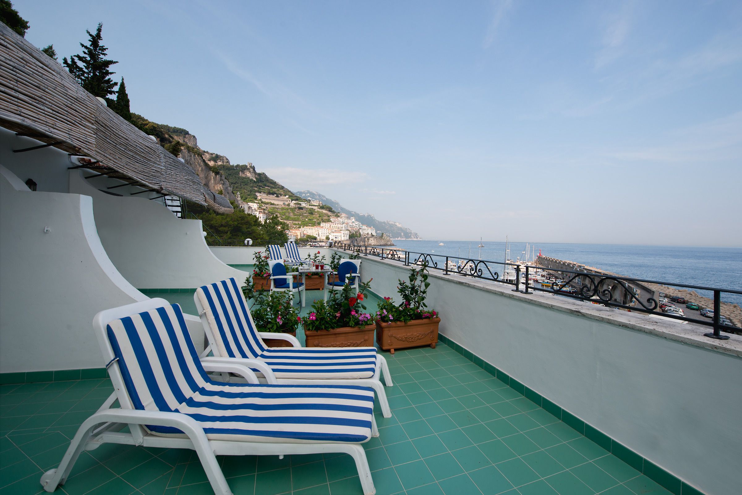 A terrace with two striped lounge chairs, a table, and potted plants. The terrace overlooks a coastal area with buildings and a clear sky.